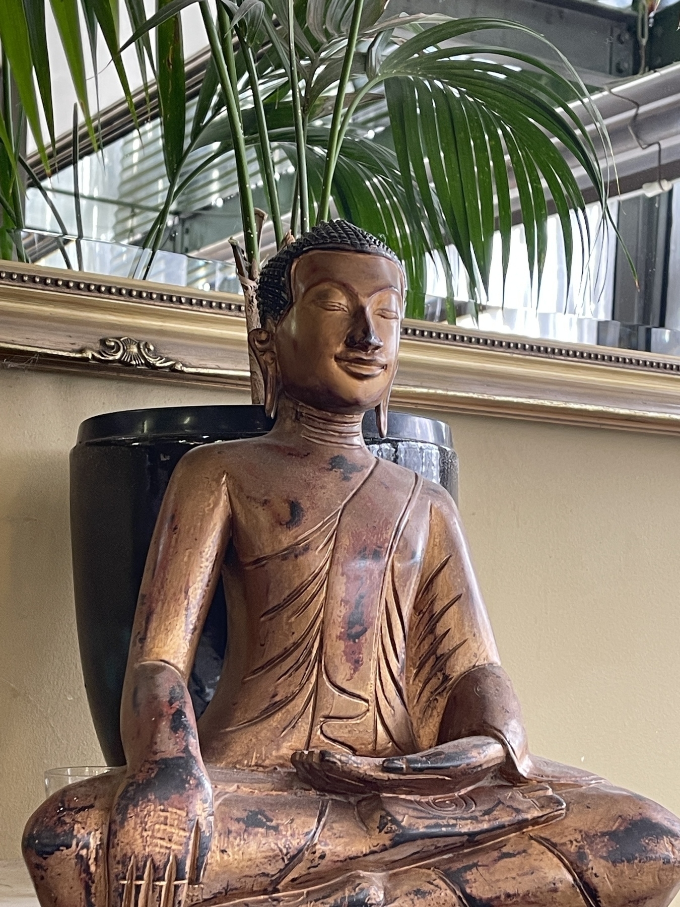 A bronze Buddha statue in a sitting position with a green palm plant behind. 